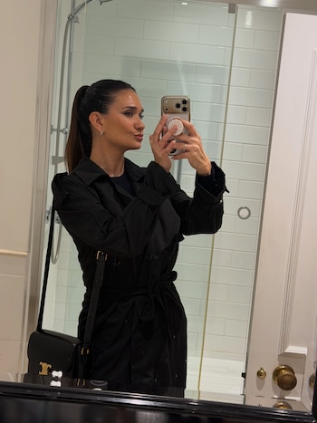 Una mujer vestida con abrigo negro y bolso sostiene un teléfono frente a un espejo en un baño con mampara de cristal