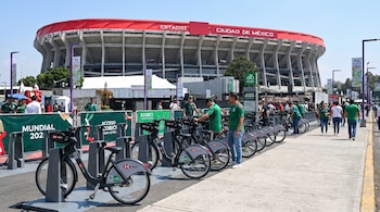 Vista frontal del Estadio Azteca con una estación de bicicletas Ecobici en primer plano, ocupada por varias bicicletas y personas a su alrededor.