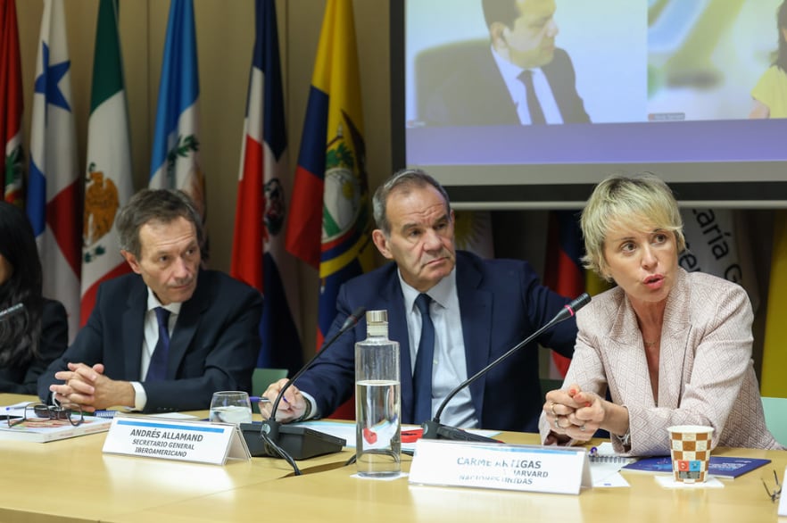 Andrés Allamand, Secretario General Iberoamericano, y Carme Artigas, de Harvard y Naciones Unidas, participan en una conferencia internacional con banderas de países de la región.