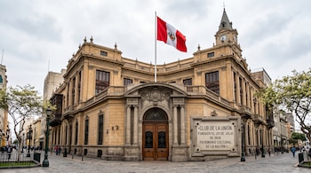 Edificio de piedra ocre con la bandera peruana en el techo, una torre con reloj y una placa grabada junto a la puerta principal.