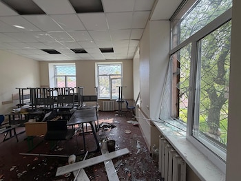 Interior de un aula con ventanas rotas, cristales en el suelo, mesas y sillas volteadas. El techo suspendido presenta paneles faltantes