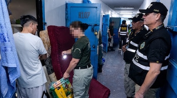 Agentes penitenciarios y un recluso en el pasillo de una prisión junto a puertas metálicas azules y colchones apoyados en la pared.
