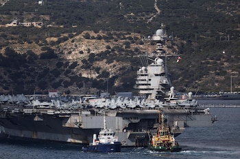 Portaaviones USS Gerald R. Ford, gris, asistido por dos remolcadores en aguas azules, con la cubierta llena de aviones militares y una costa montañosa al fondo