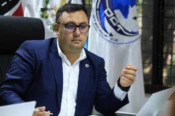 Hombre con traje azul y camisa blanca sentado en un escritorio, con gafas, laptop y bandera de DGT Guatemala de fondo