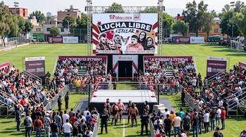 Un ring de lucha libre y una pantalla gigante instalados en una cancha de fútbol de césped rodeada por graderías con público.