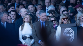 Varias personas en primera fila; hombre con traje gris sonriendo, mujer con cuello alto blanco. Detrás, una multitud. Podio oscuro con logo SRA