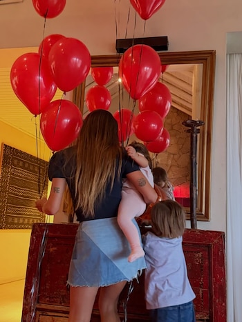Una mujer de espaldas sostiene a un niño mientras otro niño se asoma junto a un mueble rojo, rodeados de numerosos globos rojos y reflejos en un espejo