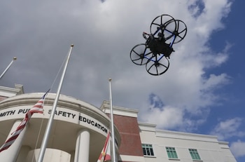 Un dron negro de cuatro hélices vuela sobre un edificio con columnas blancas, una bandera de Estados Unidos y una bandera de Florida, bajo un cielo nublado