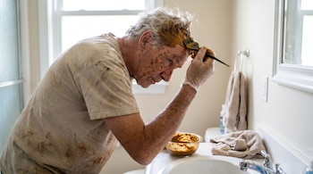 Hombre canoso aplicando henna naranja en su cabello con un cepillo en un baño. Lleva guantes blancos y tiene manchas de henna en la cara y la camiseta.