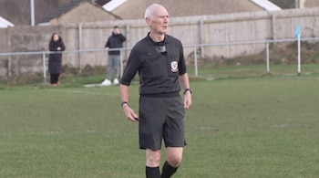 Árbitro de fútbol calvo de edad avanzada con uniforme negro en un campo de césped. Al fondo, dos espectadores, una valla de madera y una bandera azul.
