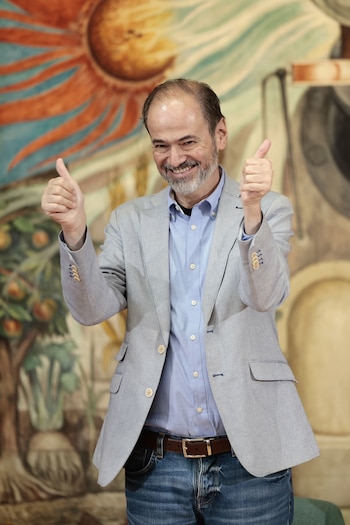 Un hombre con barba, chaqueta clara y camisa azul sonríe con los pulgares hacia arriba ante un mural ilustrado