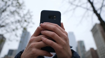 Primer plano de dos manos que sostienen un teléfono móvil oscuro, con su cámara trasera visible, y un fondo difuminado de árboles y edificios altos.