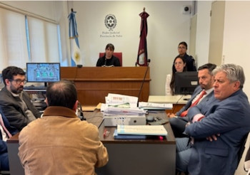 Varias personas sentadas en una sala judicial. Detrás, una persona en un estrado, banderas, un monitor, y el escudo del Poder Judicial de Salta