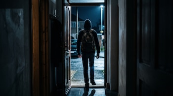 Silueta de espaldas de una persona con capucha y mochila en la puerta abierta de una casa iluminada desde la calle.
