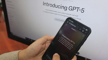 Una mano sostiene un smartphone con la interfaz de GPT-5, frente a un monitor de computadora que muestra información sobre GPT-5