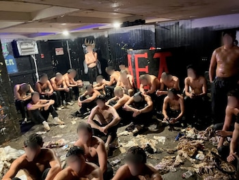 Un grupo de jóvenes sin camiseta y con el rostro difuminado permanece sentado en el suelo de un sótano cubierto de basura
