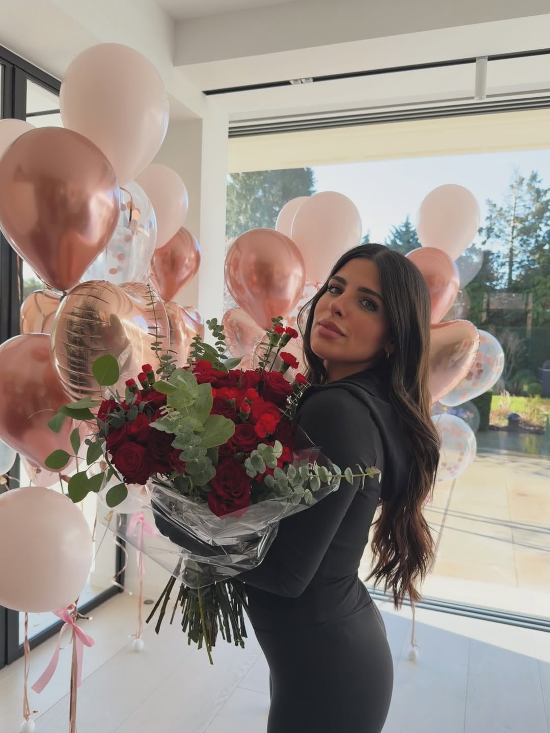 Valentina posa con un exuberante ramo de rosas rojas y eucalipto, rodeada de globos en tonos rosa y oro, celebrando un momento especial (Instagram: valucervantes)