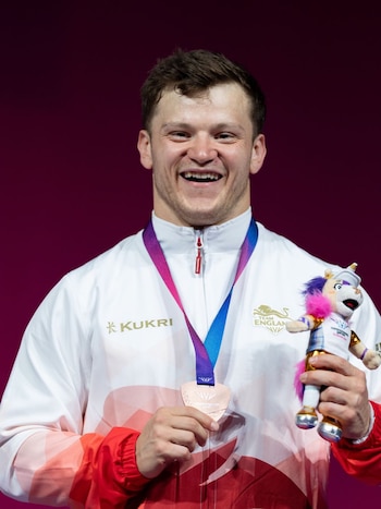 Hombre sonriendo con chaqueta blanca, medalla de bronce al cuello y mascota de unicornio. Fondo magenta. Logotipos de "Team England" y "Kukri" en la chaqueta