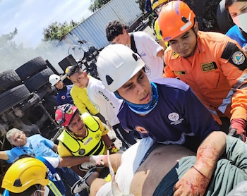 Varios socorristas con uniformes blancos y rojos inspeccionan un automóvil negro destruido junto a la estructura de un camión