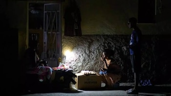Imagen oscura con tres personas iluminadas por una bombilla, una mujer agachada junto a una caja de huevos y dos hombres. Pared texturizada de fondo