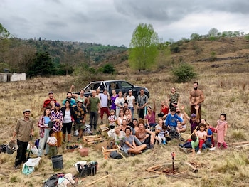 Decenas de personas de varias edades posan al aire libre en el campo con plantas en cajas de madera, herramientas y una camioneta negra