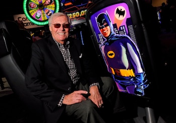 Adam West, un hombre mayor con traje y gafas de sol, sonríe sentado junto a una máquina tragamonedas de casino que muestra una imagen de Batman
