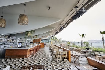 Interior de Mangos Restaurante con barra, suelo de mosaico y lámparas de mimbre; una terraza con mesas de madera y vista al mar y palmeras