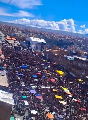 Vista aérea de una gran congregación de personas con toldos multicolores en una zona urbana, con edificios, montañas y cielo azul con nubes