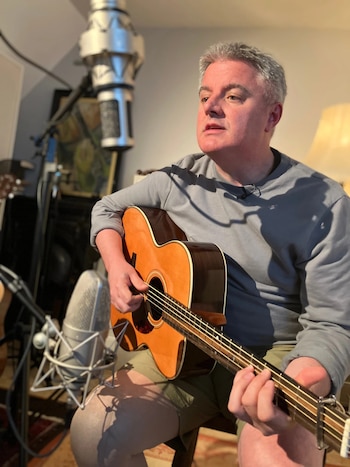 Hombre de cabello gris con camisa gris y pantalones cortos caqui tocando una guitarra acústica de madera clara, con un gran micrófono de estudio plateado en primer plano
