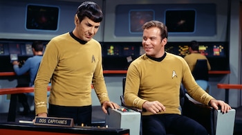 Leonard Nimoy de pie y William Shatner sentado lucen uniformes amarillos frente a una placa con el texto DOS CAPITANES.