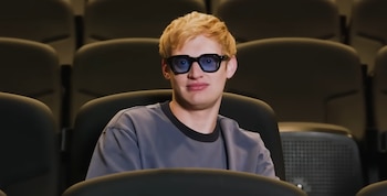 Un hombre joven con cabello rubio y gafas de montura negra y lentes azules está sentado en un asiento de cine oscuro, mirando al frente