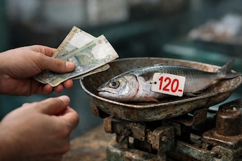 Mano sostiene dos billetes de soles peruanos. Sobre una balanza oxidada, un pescado con etiqueta de precio 120. Fondo desenfocado de mercado.