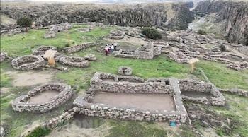 Vista aérea de las ruinas de piedra de T'aqrachullo en una colina verde. Estructuras incas, un cañón con río y personas explorando son visibles en el paisaje montañoso