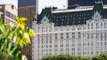Fachada de estilo clásico del hotel The Plaza con tejado verde, rascacielos al fondo y hojas en el primer plano