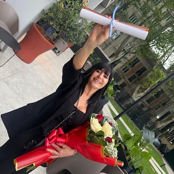Una mujer sonriente sostiene un diploma enrollado con una cinta en su mano levantada y lleva un ramo de flores rojas y blancas