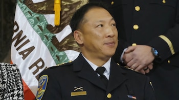 Primer plano de Derrick Lew, jefe de policía de San Francisco, con uniforme oscuro e insignias doradas, mirando a la derecha. La bandera de California está detrás