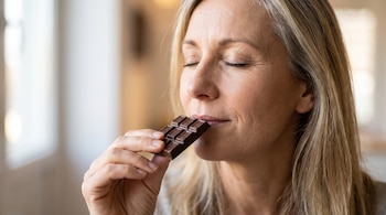 Mujer adulta con cabello rubio y canoso de ojos cerrados, lleva un trozo de chocolate oscuro a la boca con la mano.