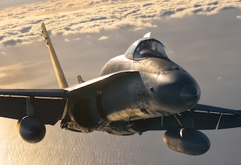 Un avión de combate gris vuela en primer plano sobre el mar y ante una capa de nubes iluminadas por el sol