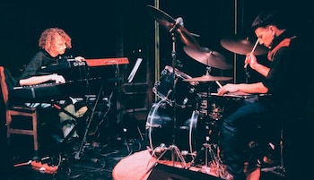 Dos músicos actúan en un escenario oscuro con iluminación roja; uno toca un teclado Casio y el otro una batería con platillos y tambores