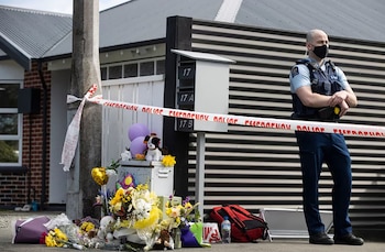 Oficial de policía con mascarilla en una calle, delante de un memorial con flores, globos y peluches, una cinta policial y buzones de correo residenciales