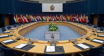 Salón de conferencias con mesa ovalada, micrófonos, carpetas, agua y letreros; al fondo, 33 banderas de diferentes países y un escudo en la pared. Documento dice "Declaración de Cusco".