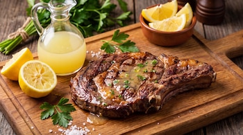Bife de costilla asado con salsa y perejil picado. Limones en mitades y gajos, una jarra con jugo. Sal y atado de perejil en tabla de madera.