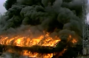 Vista aérea de un edificio grande en llamas, con intensas llamas anaranjadas a lo largo de su estructura y una columna de humo negro elevándose al cielo