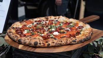 Una pizza redonda con salsa roja, rodajas de pepperoni, trozos de queso blanco y hojas de albahaca, servida sobre una pala de madera con un fondo borroso