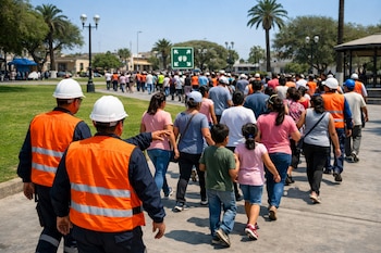 Grupo de personas, adultos y niños, en un simulacro de evacuación guiados por personal con chalecos naranja y cascos blancos en una plaza con césped y árboles