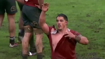 Un jugador de rugby en el suelo con la camiseta roja rota levanta la mano derecha junto a otros deportistas sobre el césped