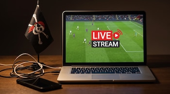 Un ordenador portátil muestra un partido de fútbol en vivo con el texto 'LIVE STREAM'. Junto a él hay una bandera pirata, un teléfono móvil y cables enredados.