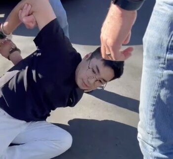 Primer plano de un hombre con gafas y camisa oscura arrodillado en el suelo, con una mano esposada y siendo sujetado por otras dos manos visibles