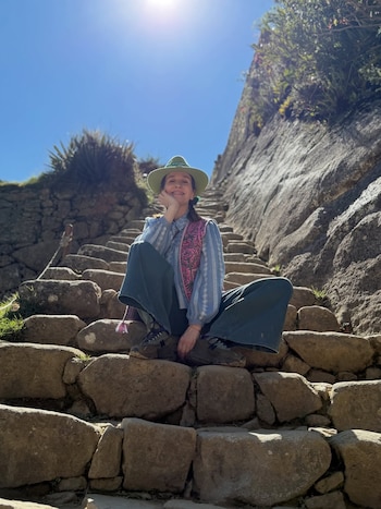 Una mujer sentada en una escalera de piedra al aire libre. Viste sombrero verde, blusa rayada, chaleco rosa y pantalones oscuros. Fondo de montañas rocosas