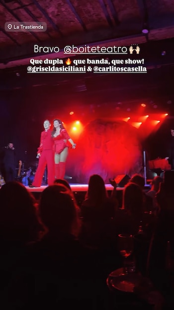 Dos personas vestidas de rojo cantan en un escenario con luces rojas frente a la silueta del público